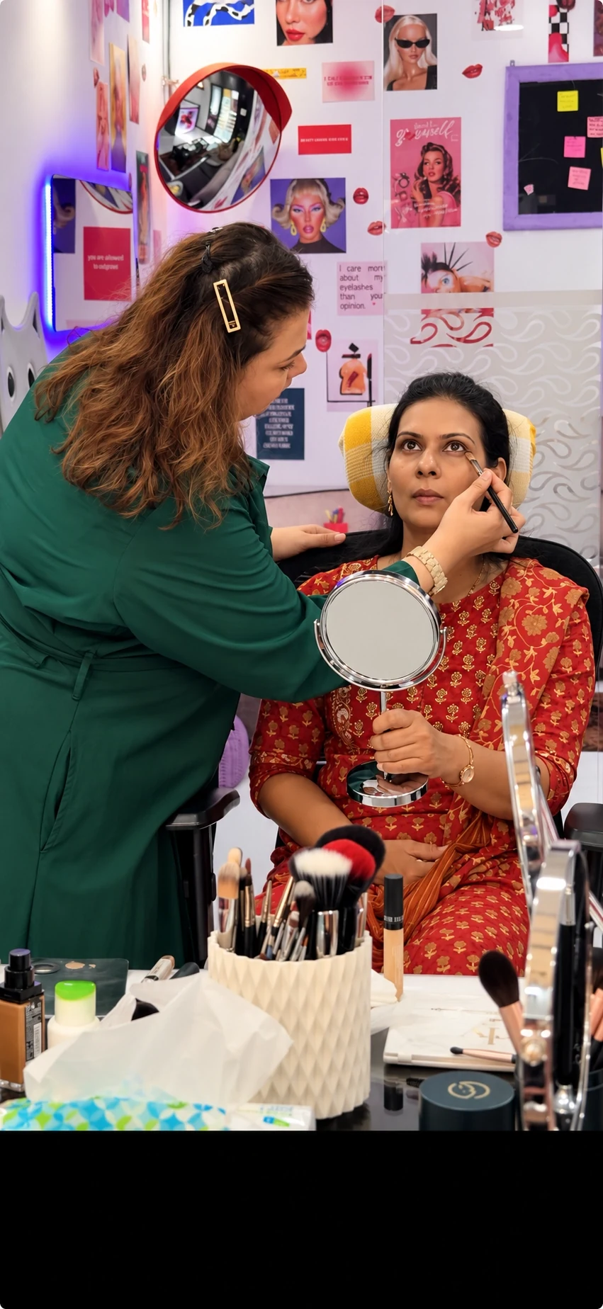 A makeup educator guiding a student through a consultation at the mirror, with skin-tone swatches and face charts behind them