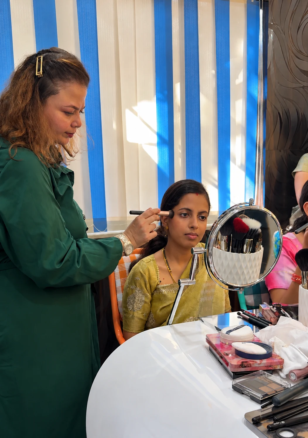 Rekha applying blush to a student seated at the vanity table